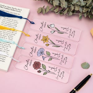 Puede incluir: Cuatro marcadores de acrílico transparente con diseños florales y el texto "Chapter Thirty" y "Sophia". Cada marcador presenta una ilustración floral diferente en tonos de blanco, amarillo, azul y rojo. También se ven un libro y un bolígrafo.