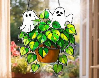 May include: A stained glass window hanging featuring two white ghost figures above a potted plant with green and yellow leaves. The plant is in a brown pot, and the window hanging is suspended by a black chain.