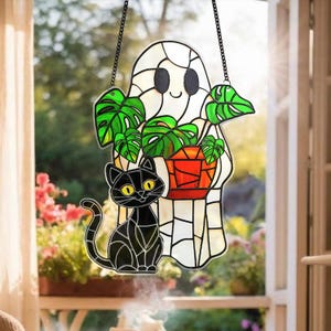 以下が含まれることがあります： ステンドグラスのハロウィーンの装飾。鉢植えの植物を持ったフレンドリーなゴーストと、下部に黒猫がいます。ゴーストは白で、黒い目と笑顔があります。猫は黄色い目をしています。緑の葉とオレンジ色の鉢も見えます。