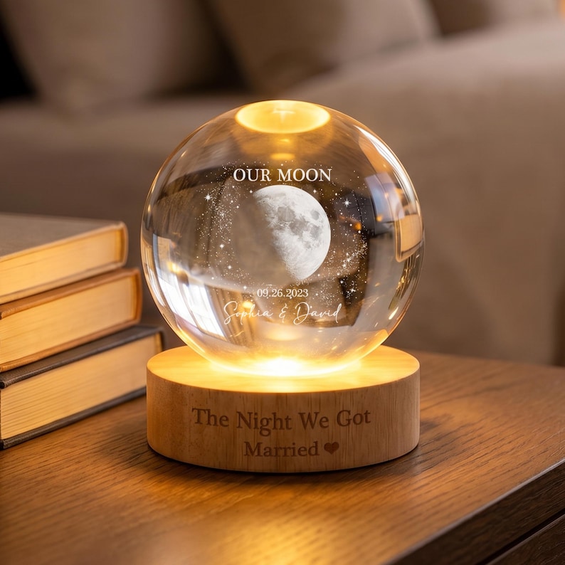 May include: A clear glass sphere lamp resting on a circular wooden base. The sphere features an etched moon, stars, the text "OUR MOON", "09.26.2023", and "Sophia & David". The wooden base is engraved with "The Night We Got Married" and a heart.