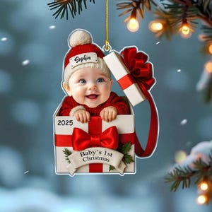 May include: A flat Christmas ornament featuring a baby in a red knit hat and sweater peeking out of a white gift box with a red ribbon. The ornament displays the name Sophia, the year 2025, and the text Baby's 1st Christmas on a banner.