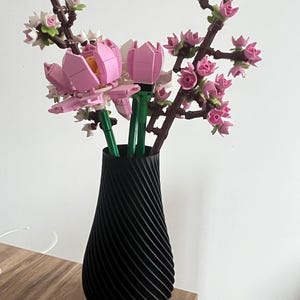 Könnte beinhalten: Eine schwarze Vase mit Spiraltextur zeigt künstliche rosa und weiße Blumen, die Kirschblüten und Tulpen ähneln. Die Vase steht auf einer Holzoberfläche vor einer weißen Wand.