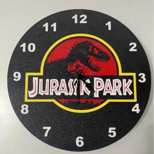 Peut inclure: Horloge murale ronde noire avec le logo Jurassic Park en rouge et jaune. Le cadran de l'horloge comporte des chiffres blancs et des aiguilles noires. Un objet décoratif pour la maison, en centimètres.