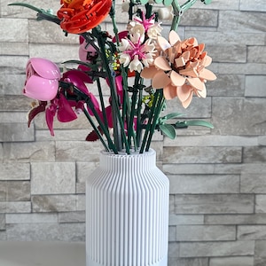 Può includere: Un vaso bianco con linee verticali contiene un colorato bouquet di fiori. I fiori sono di plastica e includono fiori arancioni, rosa e pesca, con steli e foglie verdi. Il vaso è appoggiato su una superficie bianca.
