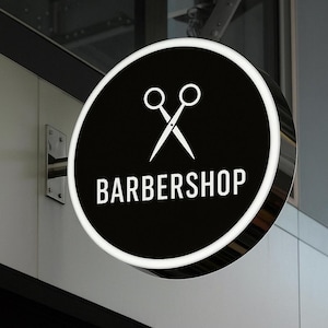 Może przedstawiać: Okrągły szyld zakładu fryzjerskiego z czarnym tłem i białymi akcentami. Znak przedstawia parę nożyczek i napis "BARBERSHOP" w białym kolorze. Znak jest zamontowany na ścianie.