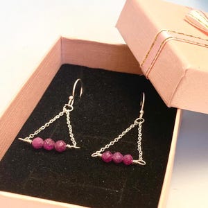 Può includere: Orecchini in argento con un design triangolare, ciascuno con tre piccole pietre preziose rosa rotonde. Gli orecchini sono esposti all'interno di un cofanetto regalo rosa chiaro aperto con interno nero.