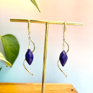 Può includere: Un paio di orecchini pendenti con gemme blu scuro sfaccettate sospese a filo argentato curvo. Gli orecchini sono esposti su un espositore per gioielli color oro, con uno sfondo sfocato di foglie verdi e un gradiente blu e rosa.