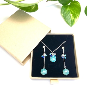 Könnte beinhalten: Ein Schmuckset mit Halskette und Ohrringen, präsentiert in einer beigen Geschenkbox. Das Set umfasst eine silberne Kette und passende Ohrringe, jeweils mit blauen und türkisfarbenen Edelsteinen verziert. Die Box ist geöffnet und zeigt den Schmuck vor schwarzem Samt.
