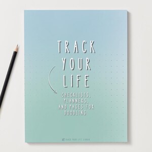 Puede incluir: Un cuaderno con una cubierta degradada azul-verde con el texto "TRACK YOUR LIFE". Incluye listas de verificación, planificadores y páginas para garabatear. Un lápiz negro con una virola dorada descansa sobre la superficie blanca.