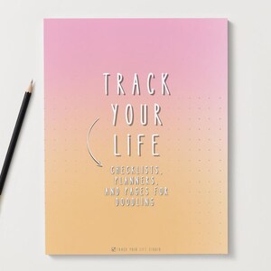 Puede incluir: Un cuaderno con un fondo degradado rosa y amarillo y el texto "TRACK YOUR LIFE". El cuaderno incluye el texto "CHECKLISTS, PLANNERS, AND PAGES FOR DOODLING". Un lápiz negro con punta dorada descansa sobre la superficie blanca.