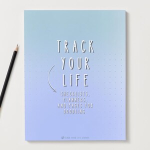Puede incluir: Un cuaderno con una cubierta degradada azul y morada y el texto "TRACK YOUR LIFE". El cuaderno incluye listas de verificación, planificadores y páginas para garabatear. Un lápiz negro con una virola dorada descansa sobre la superficie blanca.