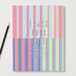 Puede incluir: Un cuaderno con un diseño de rayas coloridas en rosa, azul y melocotón. El texto "TRACK YOUR LIFE" se muestra en blanco, junto con las palabras "CHECKLISTS, PLANNERS, AND PAGES FOR DOODLING". Un lápiz negro con punta dorada descansa sobre la superficie blanca.