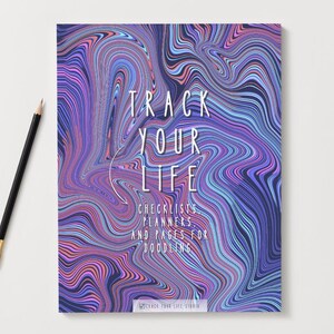 Puede incluir: Un cuaderno con un diseño abstracto y arremolinado en tonos morados, azules y rosas. El texto "TRACK YOUR LIFE" está centrado en la portada, con texto adicional debajo. Un lápiz negro con una virola dorada descansa sobre la superficie blanca.