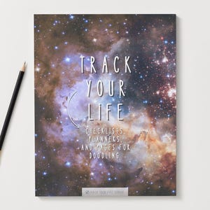 Puede incluir: Un cuaderno con un diseño de galaxia y el texto "Track Your Life". El cuaderno incluye listas de verificación, planificadores y páginas para dibujar. Un lápiz negro con punta dorada descansa sobre la superficie blanca junto al cuaderno.
