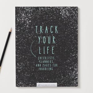 Puede incluir: Un cuaderno negro con motas blancas y el texto "TRACK YOUR LIFE" en color turquesa. Debajo, se lee "CHECKLISTS, PLANNERS, AND PAGES FOR DOODLING". Un lápiz negro con punta dorada descansa sobre la superficie blanca.