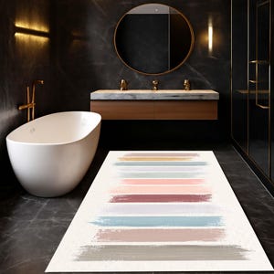 Può includere: Un tappeto da bagno rettangolare con sfondo bianco e pennellate orizzontali in vari colori, tra cui rosa, blu e grigio. Il tappeto è in un bagno moderno con vasca bianca e pareti scure.