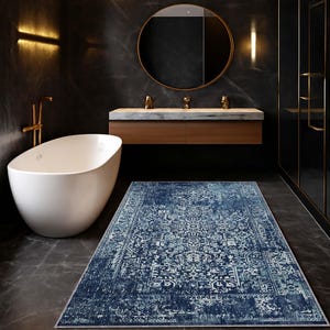 May include: A rectangular, navy blue rug with a distressed, vintage-inspired design. The rug features a detailed, faded pattern in shades of blue and white, adding a touch of elegance to the bathroom setting. The rug is placed on a dark grey tiled floor.