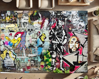 Op de afbeelding: Een kleurrijk tapijt met een graffiti-ontwerp met een cartoon eend, een soldatenachtige figuur en een portret van een persoon. Het tapijt heeft een bakstenen achtergrond met verschillende woorden en zinnen erop geschreven, waaronder "I Want Change".