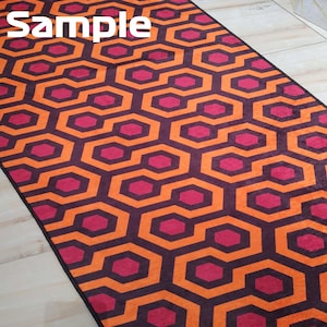 May include: A rectangular rug with a repeating geometric pattern in orange, burgundy, and dark brown. The pattern consists of interconnected hexagons and octagons. The word "Sample" is printed in white in the upper left corner.