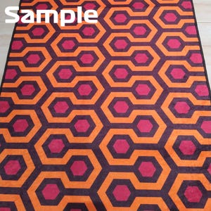 May include: A patterned rug featuring a repeating geometric design in shades of orange, purple, and magenta. The rug's pattern consists of interconnected hexagonal shapes, creating a visually striking and symmetrical pattern. The word "Sample" is printed in white in the upper left corner.