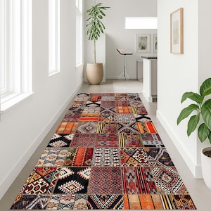 Puede incluir: Una alfombra rectangular de patchwork con varios patrones geométricos en tonos rojos, naranjas, negros y crema. La alfombra está colocada en un pasillo con paredes blancas y una planta en maceta.