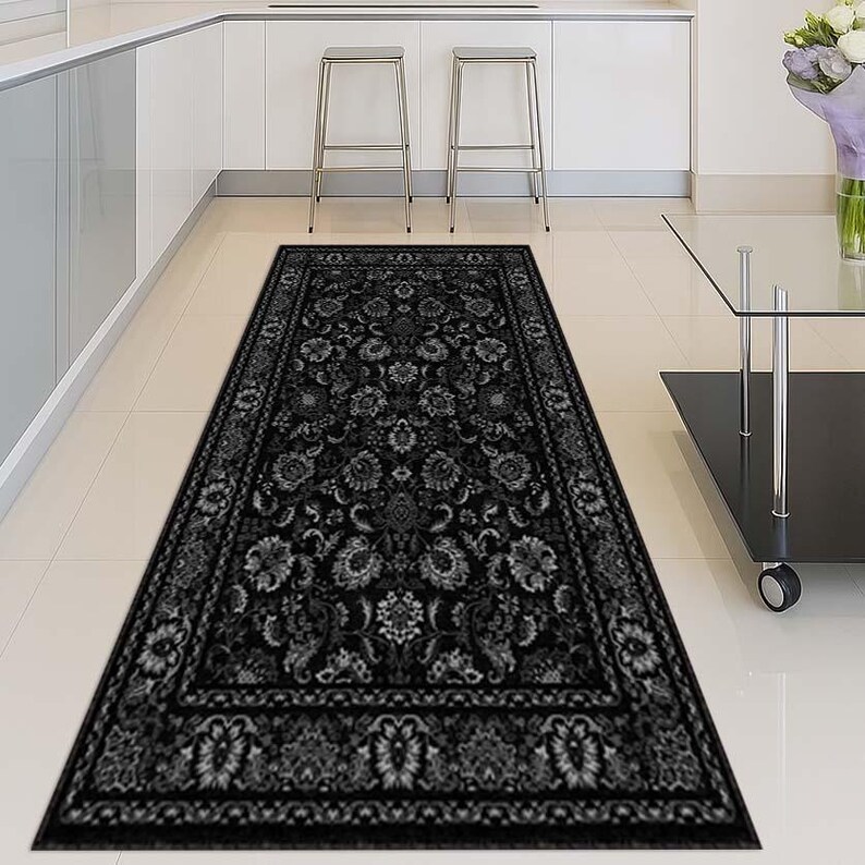 May include: A long, rectangular black rug with a detailed floral pattern. The rug features a dark border and is placed on a light-colored tiled floor. The design includes various floral and leaf motifs.