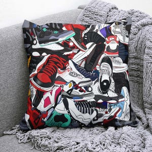 May include: Square throw pillow featuring a collage of colorful sneakers in various styles and colors, including red, black, white, and blue. The pillow is placed on a gray sofa with a matching blanket.