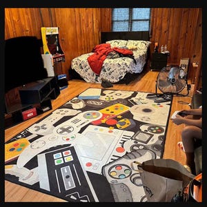 Könnte beinhalten: Ein schwarz-weißer Gaming-Teppich mit bunten Gamecontrollern und Konsolen. Der Teppich befindet sich in einem Schlafzimmer mit Holzverkleidung, einem Bett und einem kleinen Arcade-Spiel.