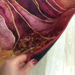May include: A close-up of a red, gold and black patterned rug with a hand holding the edge of the rug. The rug has a fringed edge.