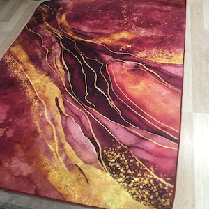 May include: Abstract area rug with a burgundy and gold colour scheme. The rug features a swirling pattern of gold lines on a burgundy background.