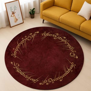 May include: A round burgundy rug with gold lettering around the edge. The lettering reads "One Ring to rule them all, One Ring to find them, One Ring to bring them all and in the darkness bind them."