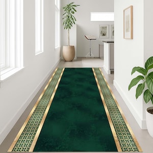 Puede incluir: Una alfombra larga y rectangular de color verde con un borde dorado y un diseño estampado. La alfombra está colocada en un pasillo con paredes blancas y una planta en maceta. Las dimensiones de la alfombra no son visibles.