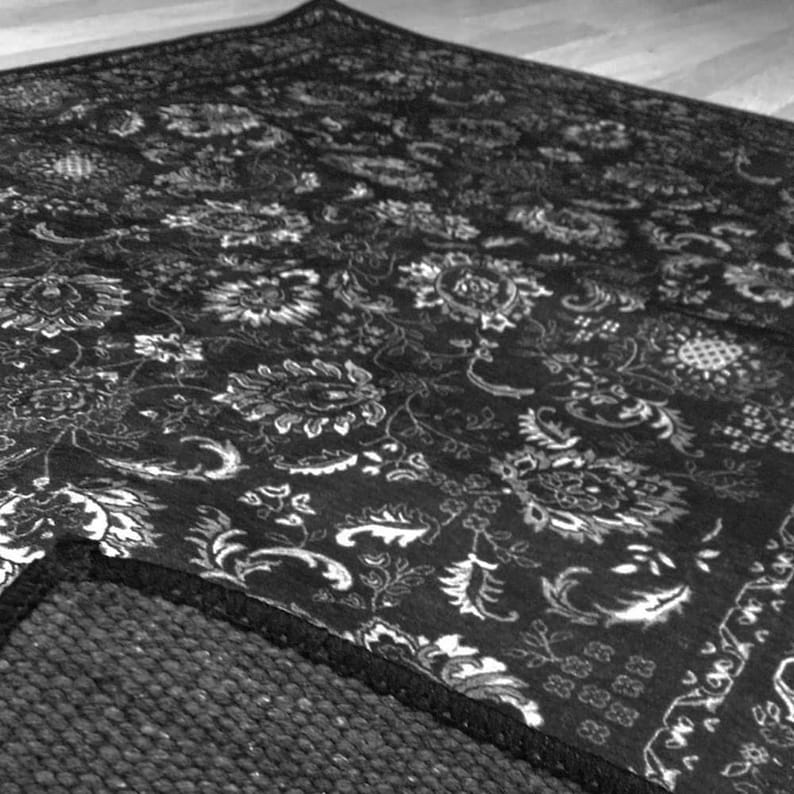 May include: Black and white floral patterned area rug. The rug features a detailed design of flowers and leaves. The rug's edge is visible, revealing a dark, textured backing. The rug is placed on a light-colored wooden floor.