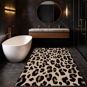 May include: A rectangular bath mat with a leopard print pattern in black, brown, and tan. The mat is placed on a dark gray tiled floor in a modern bathroom setting. A white bathtub and a wooden vanity with a round mirror are also visible.