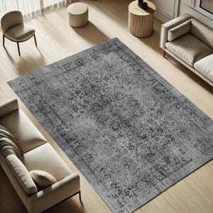 Puede incluir: Una alfombra gris rectangular grande con un intrincado diseño floral. La alfombra presenta un diseño desgastado de inspiración vintage con un borde oscuro. La alfombra está colocada sobre un suelo de madera clara, con muebles visibles en el fondo.