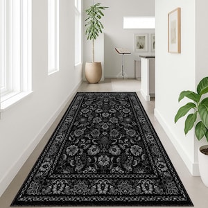 May include: A long, rectangular black rug with a floral pattern. The rug features a detailed design with various floral and leaf motifs in shades of gray and white. The rug is placed in a hallway with white walls and a potted plant.
