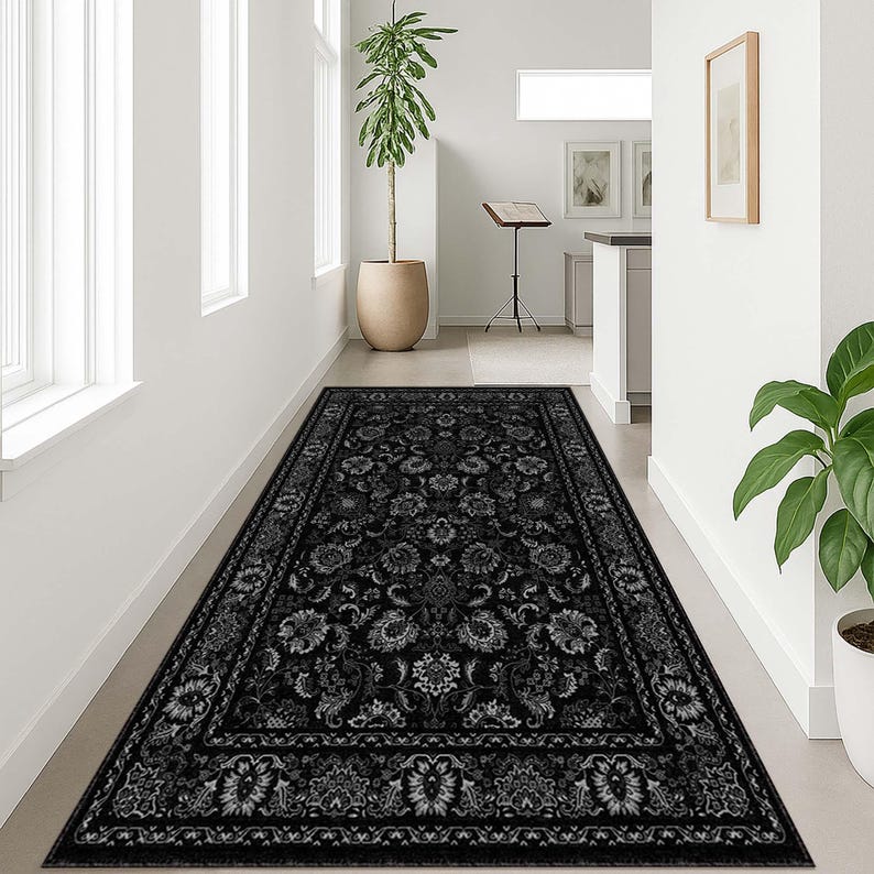 May include: A long, rectangular black rug with a floral pattern. The rug features a detailed design with various floral and leaf motifs in shades of gray and white. The rug is placed in a hallway with white walls and a potted plant.