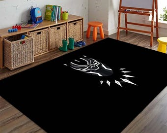 Puede incluir: Alfombra rectangular negra con un diseño gráfico en blanco y negro de una máscara de superhéroe. La alfombra está colocada sobre un suelo de madera en una habitación con muebles y juguetes infantiles. El diseño de la alfombra es llamativo.