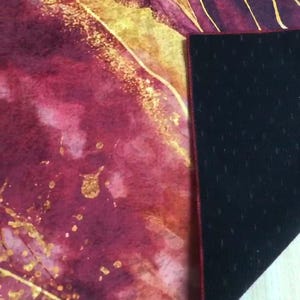 May include: A burgundy and gold watercolour patterned yoga mat with a black non-slip backing. The mat has a red stitched edge.