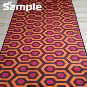 May include: A rectangular rug with a repeating geometric pattern of orange and burgundy interlocking hexagons. The rug has a black border and the word "Sample" is printed in white at the top.