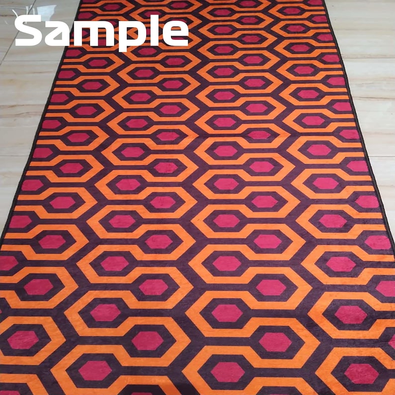 May include: A rectangular rug with a repeating geometric pattern of orange and burgundy interlocking hexagons. The rug has a black border and the word "Sample" is printed in white at the top.