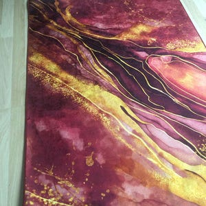 May include: Abstract art rug with a burgundy and gold colour scheme. The rug features a swirling pattern of gold lines on a burgundy background.