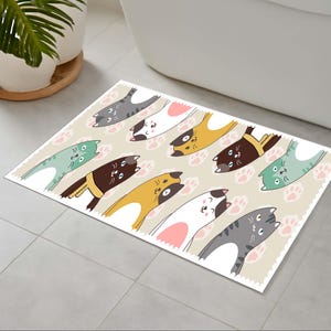 May include: A rectangular bath mat with a repeating pattern of cartoon cats in shades of green, brown, yellow, and grey. The cats are set against a beige background with pink paw prints. The mat has a scalloped edge, and measures approximately 70cm x 50cm.