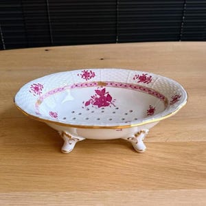 Puede incluir: Un cuenco de porcelana blanca con pie, borde dorado y detalles florales. El cuenco tiene una textura tejida alrededor del borde y está decorado con diseños florales rosas. El cuenco está sostenido por tres pequeños pies ornamentados.