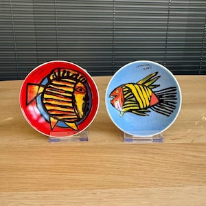Op de afbeelding: Twee decoratieve keramische borden met visontwerpen. Eén bord is rood met een gele, zwarte en blauwe vis. Het andere bord is blauw met een gele, zwarte en rode vis. Beide borden staan op transparante standaards.