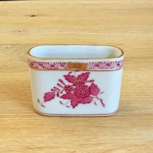 May include: A rectangular white porcelain container with a gold rim and a floral design. The container features a pink floral pattern on the front and a decorative pink and gold border around the top.