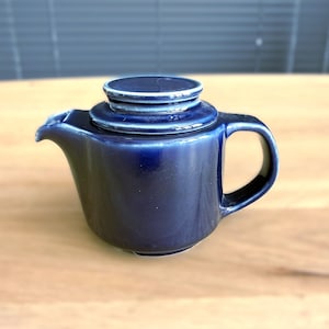 May include: A deep cobalt blue ceramic teapot featuring a glossy finish, a short curved spout, a sturdy side handle, and a matching circular lid with a stepped design. The glaze shows subtle variations in colour intensity across the rounded body.