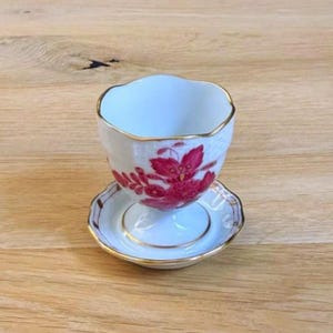 以下が含まれることがあります： 赤色の花柄が施された白いティーカップとソーサーのセット。カップとソーサーには金のトリムが付いています。ティーカップは、小さなマッチングソーサーの上にあります。セットは木製の表面にあります。