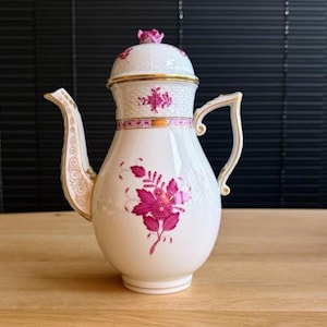 May include: A white porcelain coffee pot with a lid and handle, adorned with gold trim and a floral design in shades of pink. The pot features a curved spout and a decorative handle, set on a wooden surface.