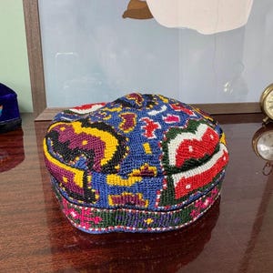 Può includere: Un cappello colorato e fantasia con una corona arrotondata. Il motivo ricamato presenta un mix di blu, rosso, giallo e viola. Il bordo è verde scuro con dettagli floreali. Il cappello è probabilmente fatto a mano.